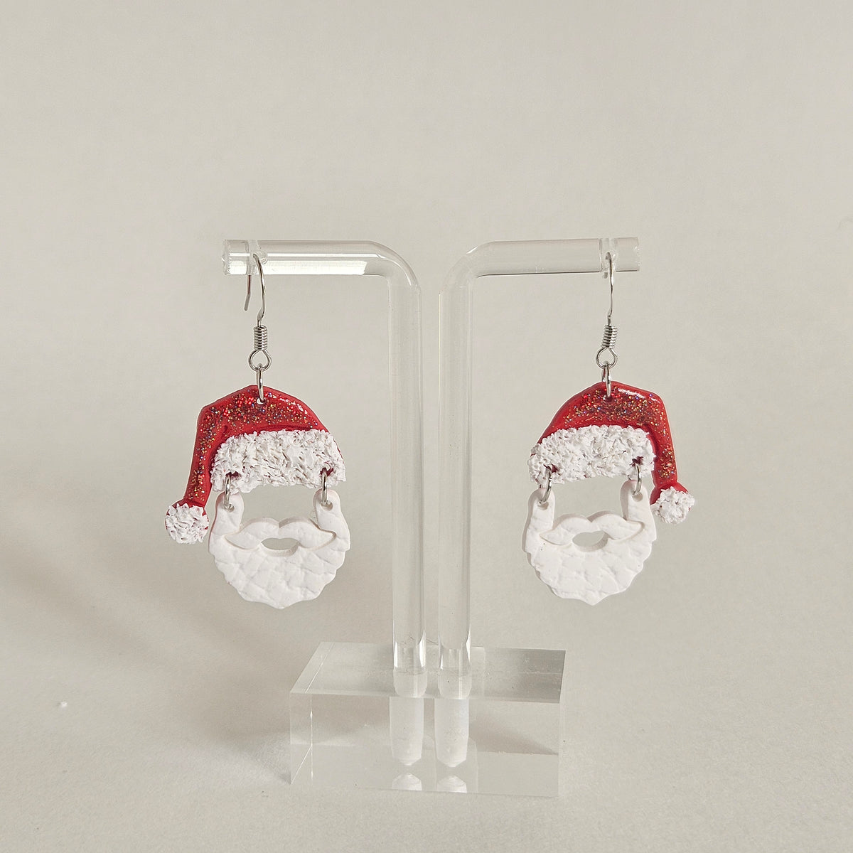 A pair of dangling earrings displayed on clear acrylic stands against a plain light background. Each earring features a red glitter Santa hat with white faux-fur trim and a white Santa beard.