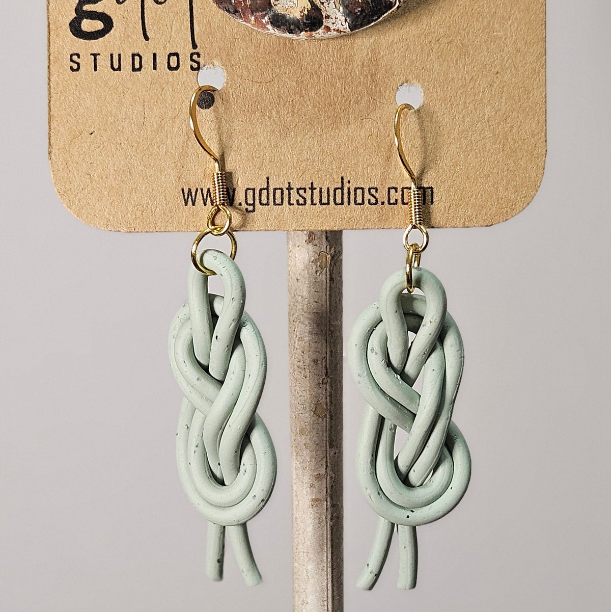 Image of handcrafted light sage bend knot macrame earrings displayed on a gdot studios card with a nuetral background.