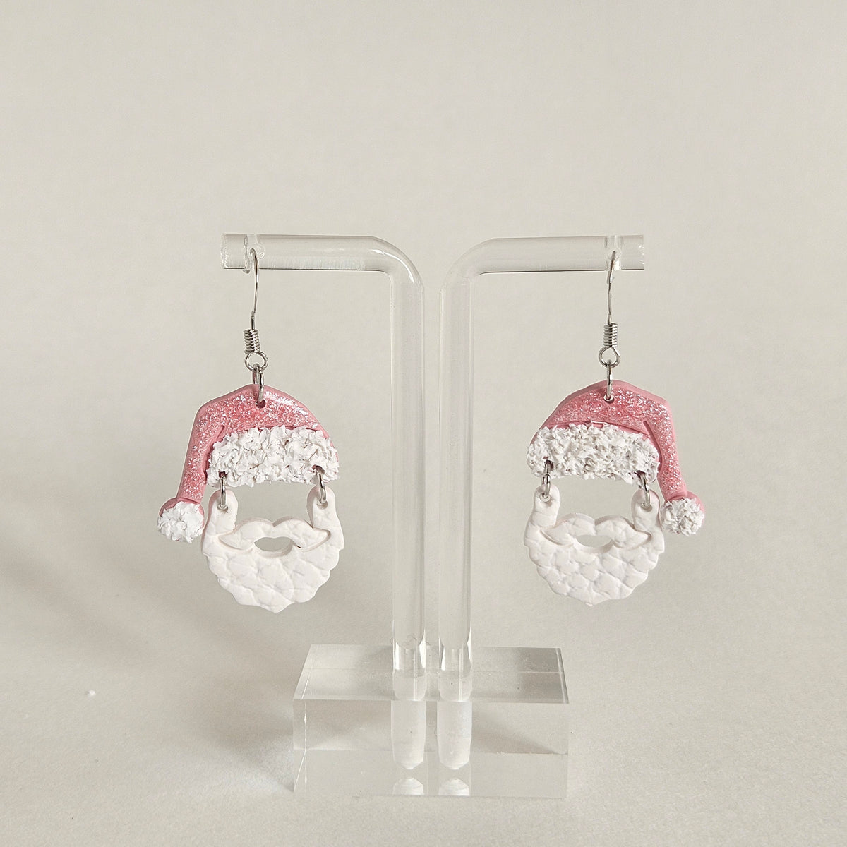 A pair of dangling earrings displayed on a clear acrylic stand against a light beige background. Each earring features a glittery pink Santa hat with white trim and a white textured beard dangle.