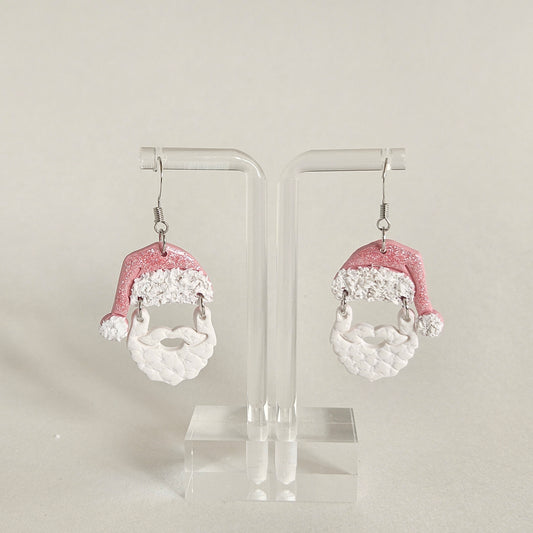 A pair of dangling earrings displayed on a clear acrylic stand against a light beige background. Each earring features a glittery pink Santa hat with white trim and a white textured beard dangle.