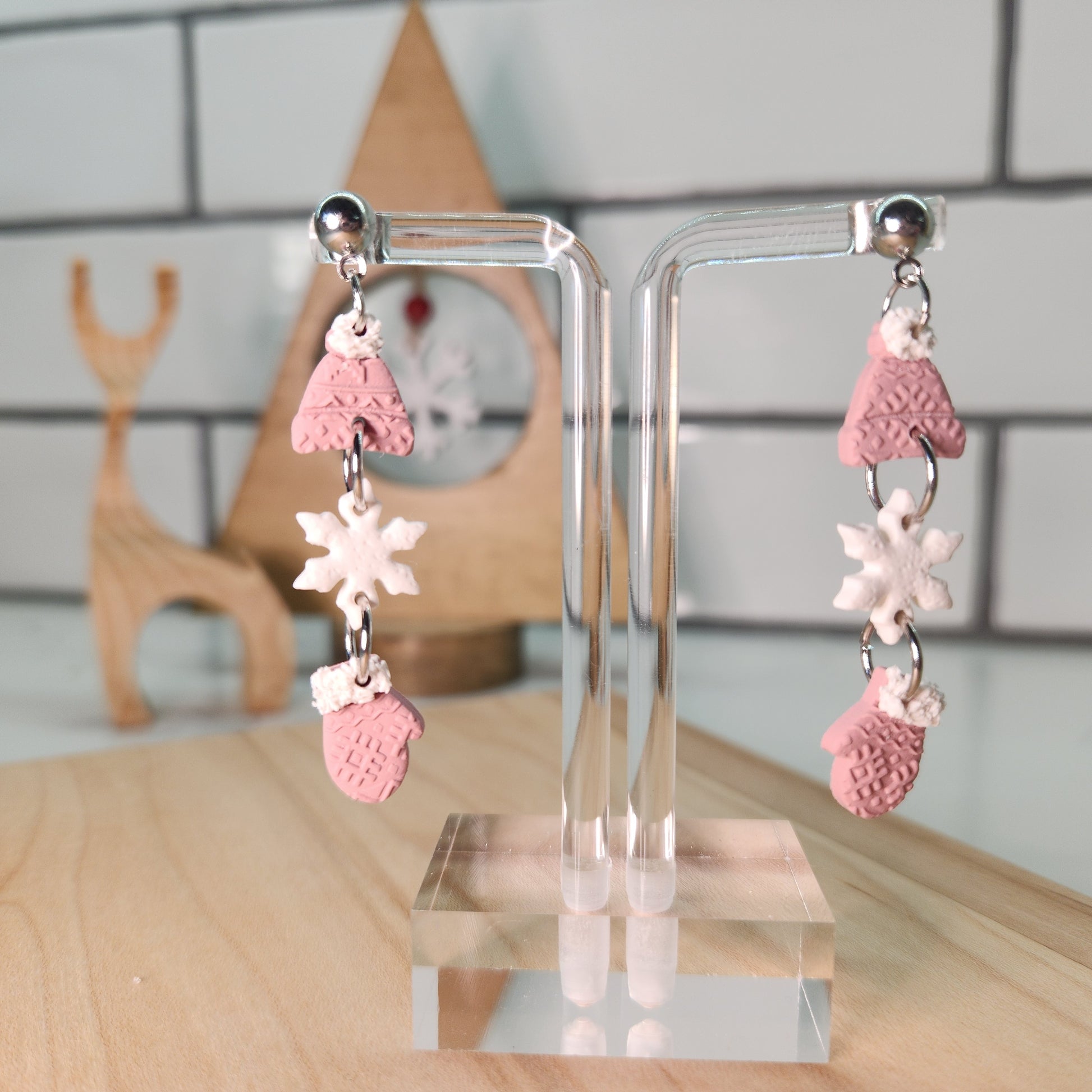 Pair of pink and white earrings making a trio of mittens, snowflakes and winter hats on a clear stand with a blurred background