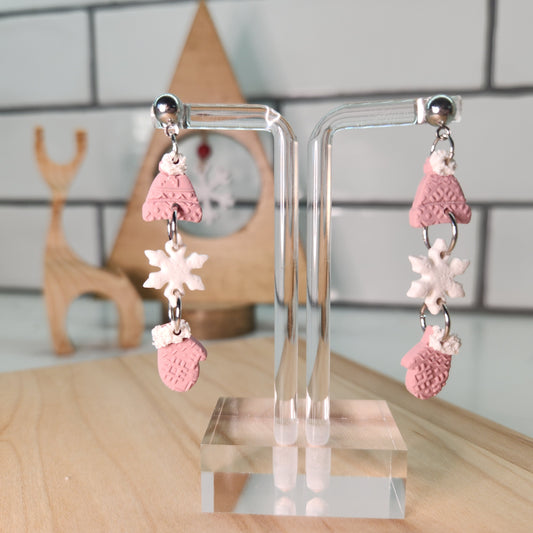 Pair of pink and white earrings making a trio of mittens, snowflakes and winter hats on a clear stand with a blurred background